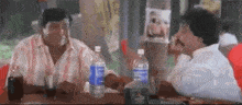 two men are sitting at a table with bottles of water and drinking coke .