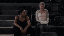 two women are sitting next to each other in a dark room and one of them is pointing at the camera .