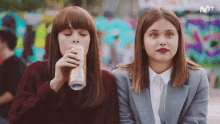 two women sitting next to each other one drinking from a can and the other wearing a suit
