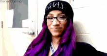 a woman with purple hair and glasses is wearing a beanie and making a funny face .