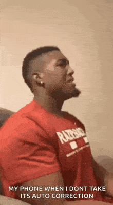 a man in a red shirt is sitting on a couch talking on his phone .