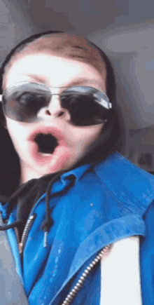 a boy wearing sunglasses and a blue jacket is making a funny face with his mouth open .