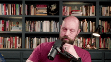 a bald man with a beard is talking into a microphone in front of a bookshelf filled with books