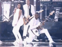 a man is playing a guitar on stage while a woman sings and a man is kneeling down .