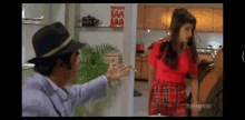 a woman in a red top and plaid skirt is standing next to a man in a black hat in a kitchen
