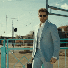 a man in a light blue suit and sunglasses stands in front of a blue gate
