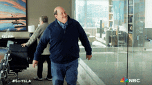 a man in a blue sweater is walking in a room with a nbc logo on the bottom right