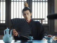 a man in a kimono is sitting at a table holding a fan in his hand .