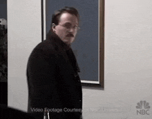 a man with glasses and a mustache is standing in front of a wall .