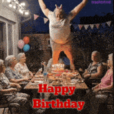 a group of elderly women sit at a table with a birthday cake and a man jumping in the air