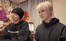 two young men are sitting next to each other in a living room . one of the men is wearing a beanie .