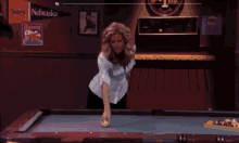 a woman stands at a pool table with a nebraska banner on the wall behind her