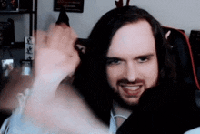 a man with long hair and a beard is sitting in a chair and waving his hand .