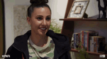 a woman wearing a camouflage shirt is smiling in front of a bookshelf with # mafs written on it