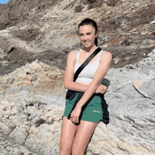 a woman wearing green shorts and a white tank top is standing on a rock .