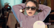 a woman wearing sunglasses and a purple sweatshirt is sitting at a table with her hands behind her head .