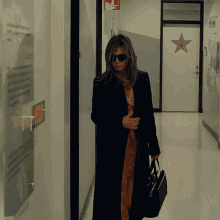 a woman wearing sunglasses and a black coat walks down a hallway with a star on the door
