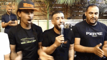 a man in a perre shirt sings into a microphone while two other men look on