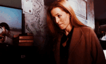 a woman in a brown coat stands in front of a stack of books and a tv