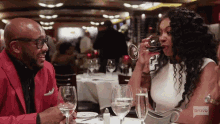 a man in a red jacket sits next to a woman drinking wine in a restaurant with bravo written on the screen