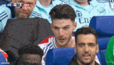 a group of men are sitting in a stadium watching a soccer game and one of them is wearing an emirates shirt