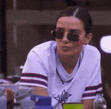 a woman wearing sunglasses and a white shirt is sitting at a table with a water bottle .