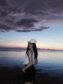 a woman wearing a white hat stands in front of a body of water at sunset