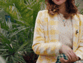 a woman wearing a yellow and white plaid jacket is standing in front of a plant