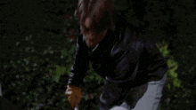 a man in a black jacket and white pants is standing in the grass at night .
