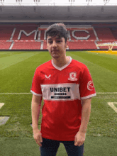 a young man wearing a red unibet jersey stands in front of a soccer field