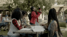 a group of people are sitting around a picnic table and one of them is wearing a red bomber jacket