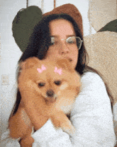a woman wearing glasses is holding a small brown dog with pink bows on its face