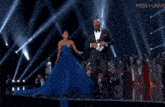 a man in a tuxedo is holding a microphone while a woman in a blue dress holds her hand out to him