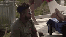 a man wearing sunglasses is sitting on a deck looking at a woman 's feet .