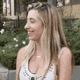 a woman in a white tank top is smiling in front of a garden