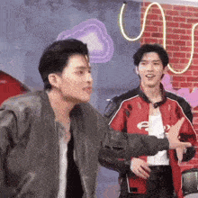 two young men are standing next to each other in a room . one of the men is wearing a leather jacket .