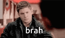 a man in a black jacket is sitting at a table with the word brah on the screen .