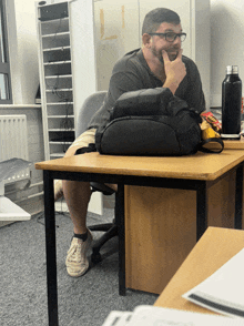 a man with glasses sits at a desk with a backpack on it
