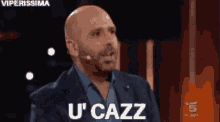a bald man in a suit and blue shirt is talking into a microphone and saying u ' cazz .