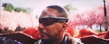 a man wearing sunglasses and a leather jacket is standing in front of a pink background .