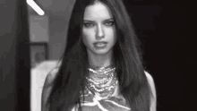 a black and white photo of a woman with long hair wearing a necklace and looking at the camera .