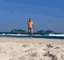 a shirtless man in blue swim trunks walks barefoot on the beach