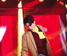 a man in a tan jacket and a yellow turtleneck is standing in front of a red wall