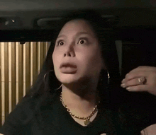 a woman wearing a black shirt and a gold necklace is making a funny face while sitting in a car .