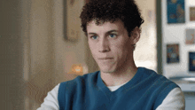 a young man with curly hair wears a blue vest