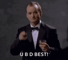 a man in a tuxedo is holding a glass of wine and saying `` ubd best '' .