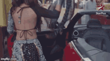 a woman in a backless top is standing next to a red car with a star plus logo