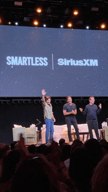 three men are standing on a stage in front of a large screen that says smartless siriusxm