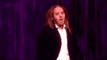 a man in a tuxedo stands on a stage in front of a purple curtain with a bbc logo on it