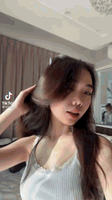 a woman in a white tank top is standing in front of a mirror and holding her hair .
